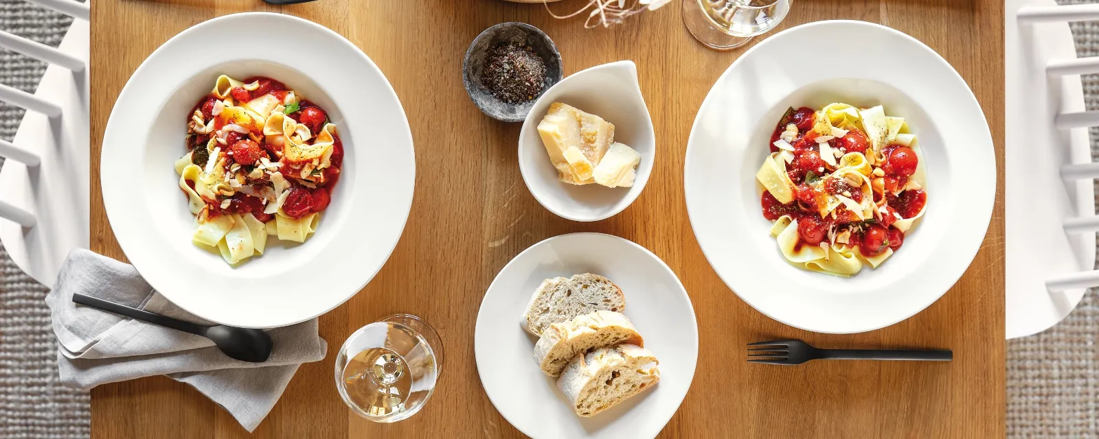 Twee Flow borden met pasta, tomaten en kaas van Villeroy & Boch op een houten tafel, vergezeld van een schaal brood, twee wijnglazen, bestek en een decoratief middenstuk.