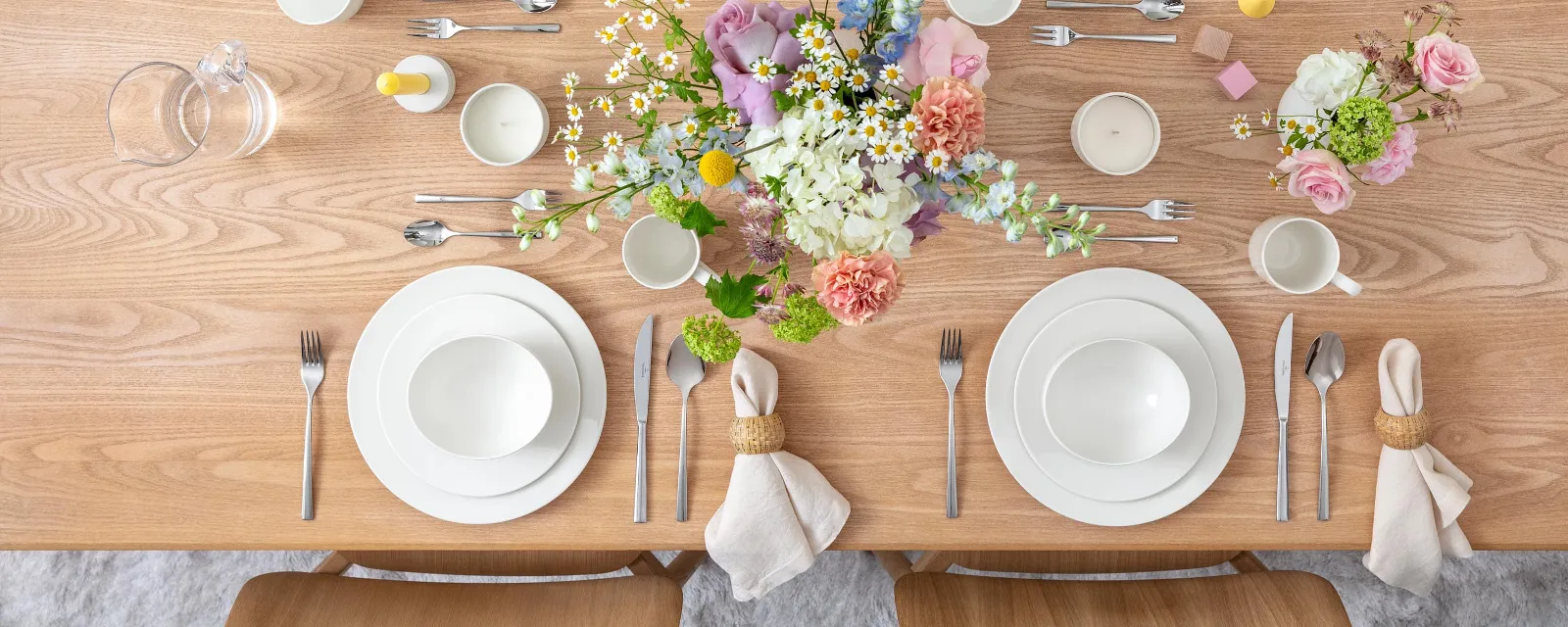 A wooden dining table set for four people with white plates, cups, and cutlery. In the centre, there is a floral arrangement with pink and white flowers. Glass jugs are ready, and rolled napkins are placed next to the plates.