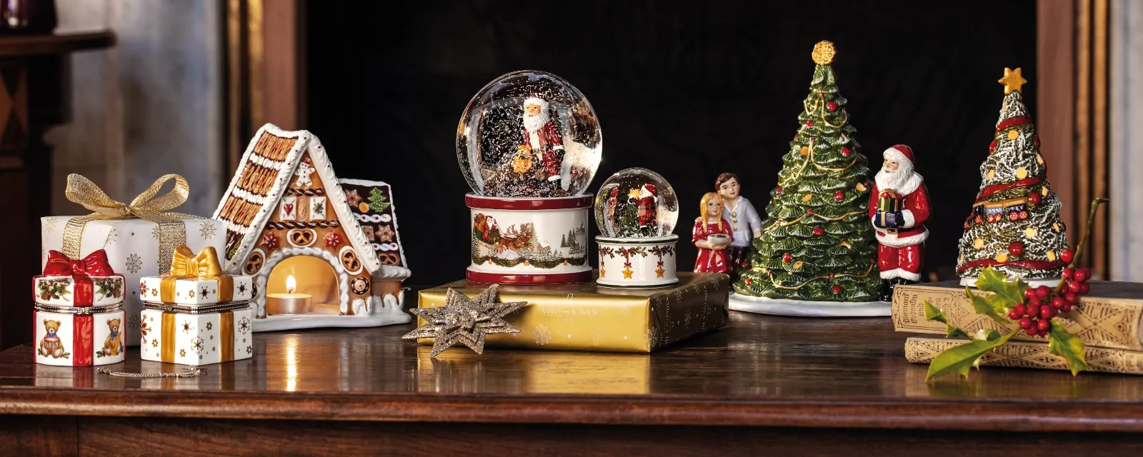 Une scène chaleureuse au coin du feu avec une couronne, des boules de neige du Père Noël, une figurine de sapin de Noël, une maison en pain d’épice, des cadeaux et le jouet en forme de voiture « A Christmas Toy » de Villeroy & Boch sur un rebord de cheminée en bois.