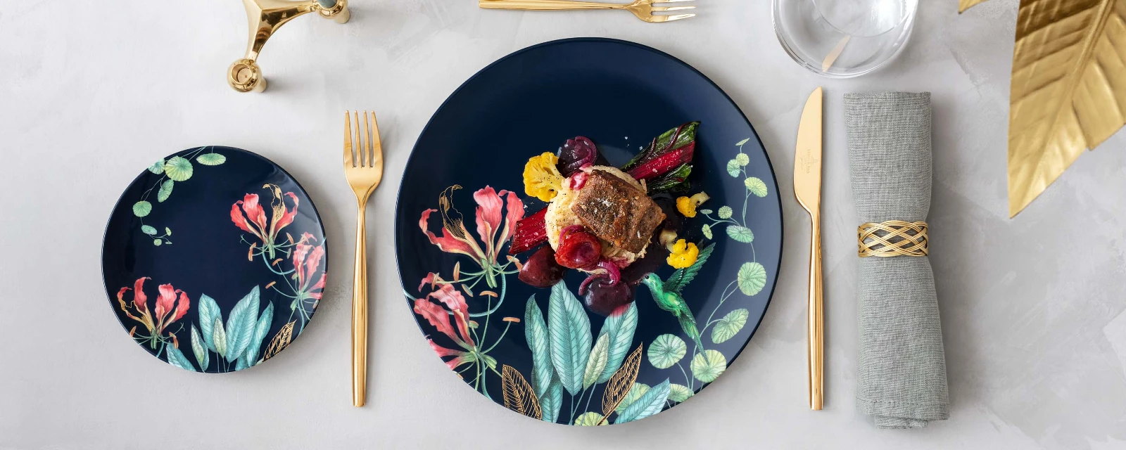 Elegant dining table with Avarua plates and a gourmet dish, accompanied by golden MetroChic flatware and a glass of red wine.