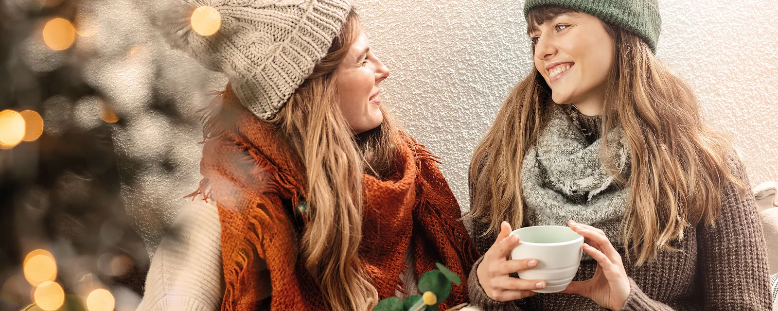 Twee vrouwen in winterkleding glimlachen naar elkaar terwijl ze gaan zitten. De een houdt een cadeau vast, de ander een It's My Match-kopje van Villeroy & Boch. Twee vrouwen in winterkleding glimlachen naar elkaar terwijl ze gaan zitten. De een houdt een cadeau vast, de ander een It's My Match-kopje van Villeroy & Boch. Op de voorgrond is vaag een kerstboom te zien.
