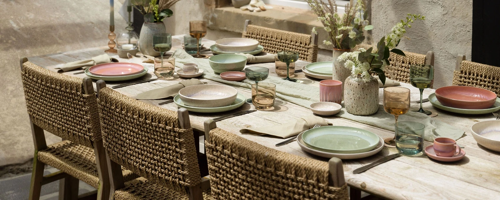 Une présentation de table de salle à manger rustique pour l’extérieur avec des chaises tressées, des assiettes Perlemor de couleur pastel like. by Villeroy & Boch, des verres et des centres de table floraux sur un fond en pierre avec une fenêtre.