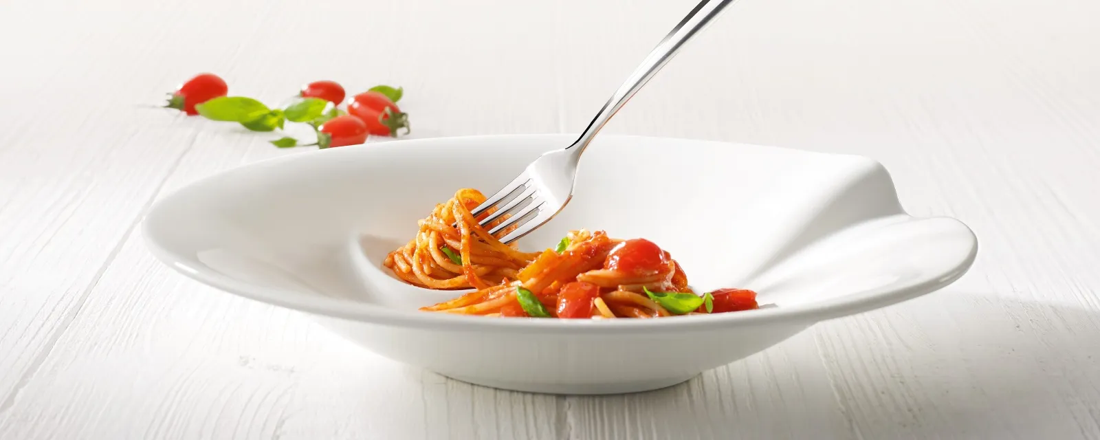 A fork in a white Villeroy & Boch Pasta Passion bowl holds a small portion of spaghetti with cherry tomatoes and basil on a light-colored wooden table.