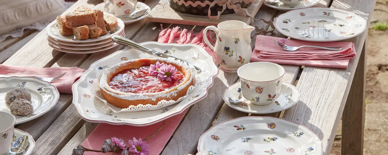 The Petite Fleur collection from Villeroy & Boch is elegantly arranged on a wooden table and includes plates with a floral pattern, a cake, a tart, cups, and pink napkins for tea.