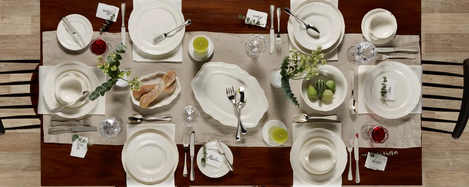 A top view of a wooden dining table set for six people with white crockery from the Villeroy & Boch Manoir collection, along with cutlery, glassware, green accents, bread and place cards on a beige table runner.