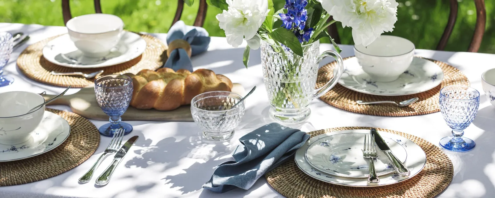 Een tuintafel is gedekt met "Vieux Luxembourg" bloemenporselein van Villeroy & Boch, rieten placemats, blauw getint glazen servies, een kristallen kan met blauwe en witte bloemen en een brood.