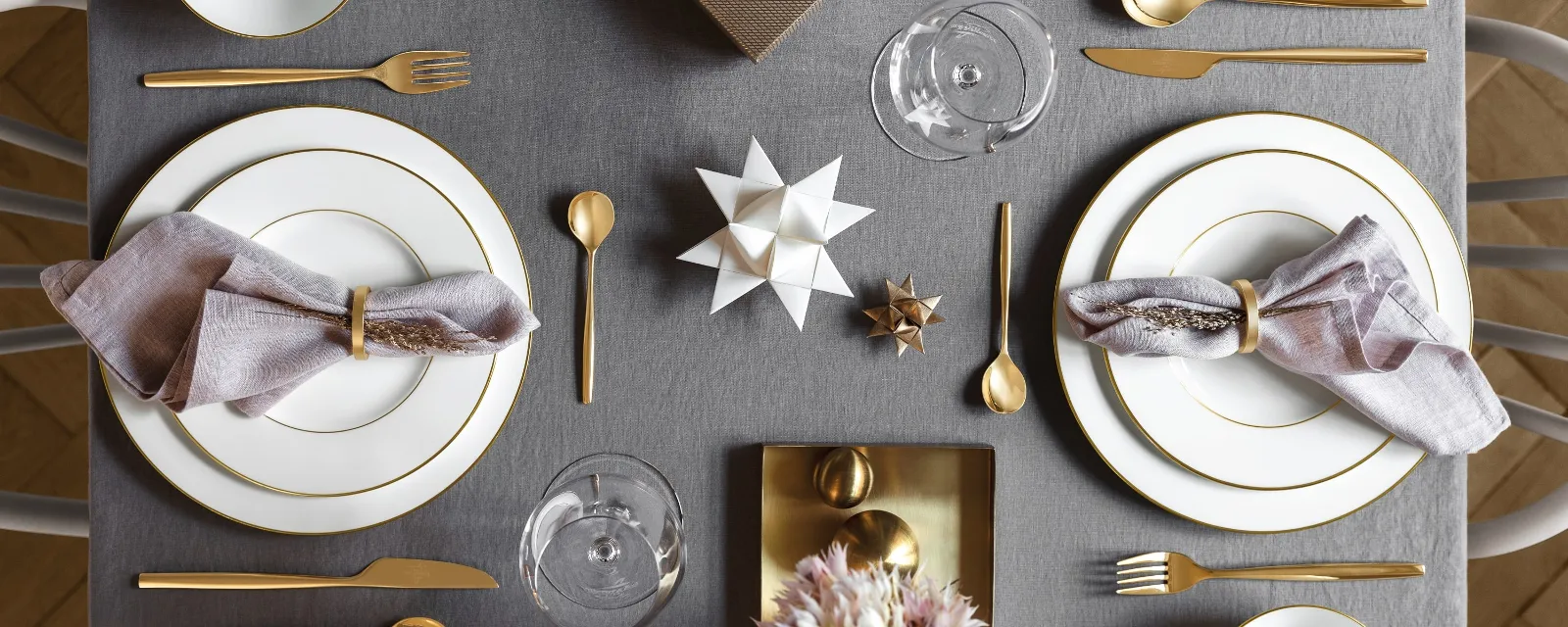 Een elegant gedekte eettafel met twee Anmut couverts van Villeroy & Boch, gouden bestek, lavendelkleurige servetten, een geschenkdoos en een bloemstuk op een grijs tafelkleed.