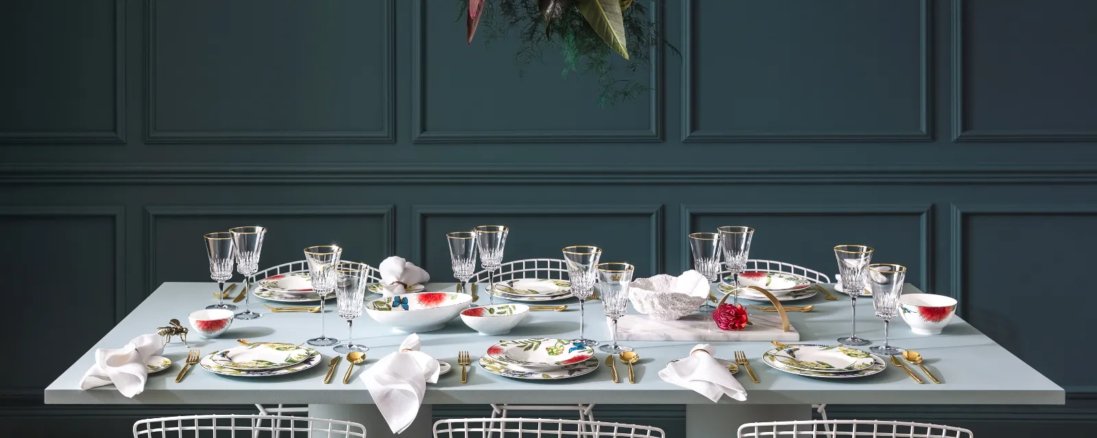 Le set de table Amazonia de Villeroy & Boch se compose de verres, de couverts de table dorés et d’assiettes aux motifs floraux. Une composition florale est suspendue au-dessus de la table de la salle à manger, ce qui crée un joli contraste avec le mur aux lambris sombres.