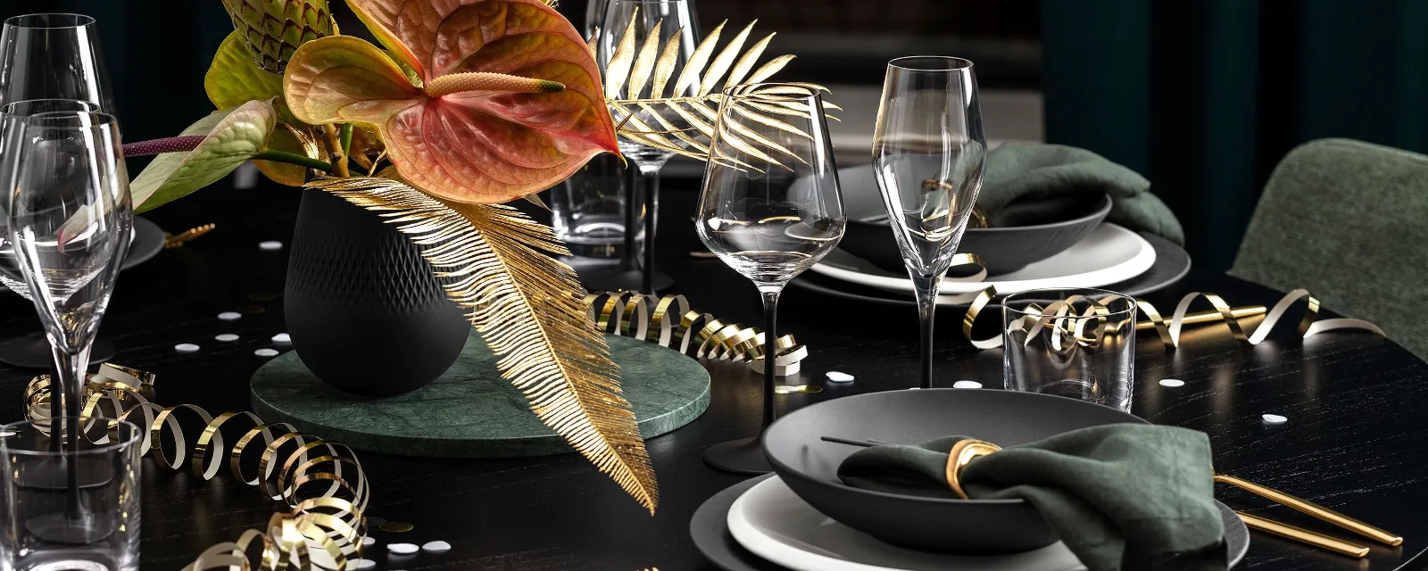 Silvester Mix & Match mit NewMoon und Manufacture von Villeroy & Boch Elegantes Tischgedeck von Villeroy & Boch mit dunklen Manufacture Tellern, goldenem Besteck, grünen Servietten und einer schwarzen Collier Vase mit tropischen Blumen und Blättern. Goldene Luftschlangen und Gläser schmücken den schwarzen Tisch.