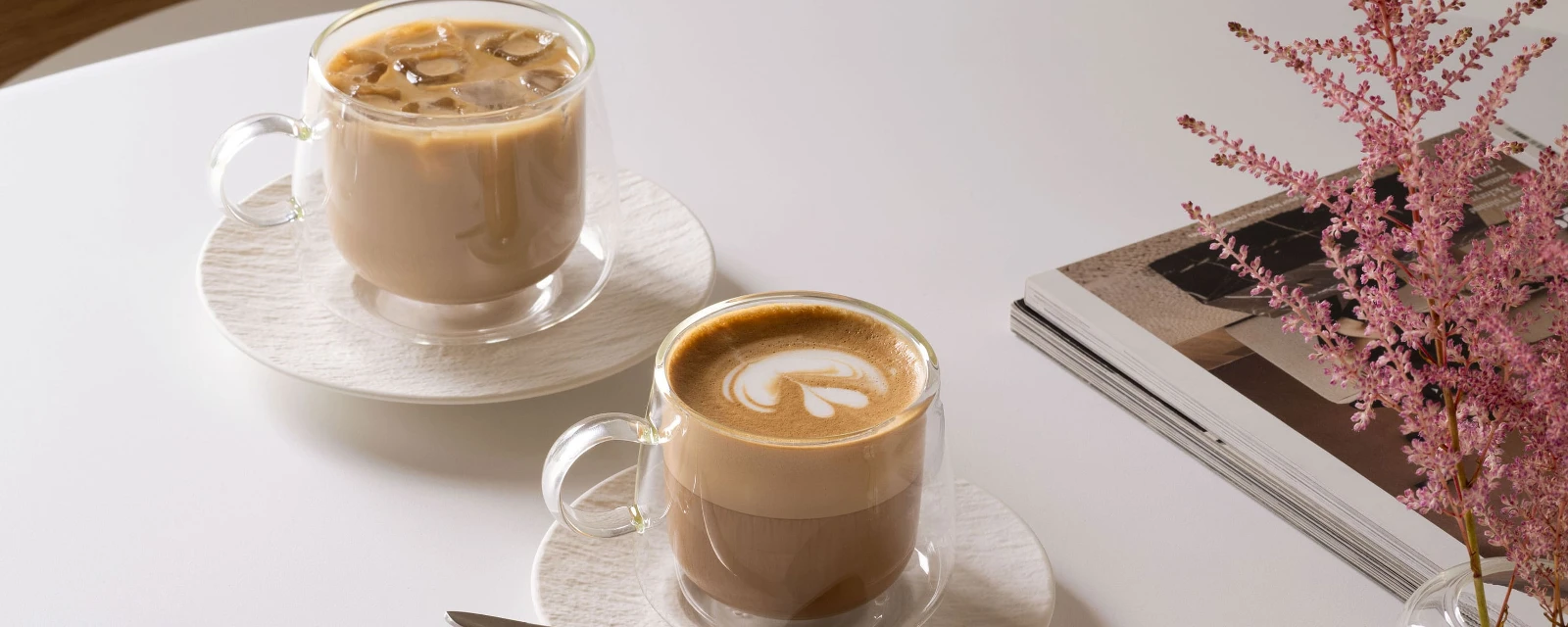 Twee glazen Villeroy & Boch Artesano-bekers op een tafel: de ene gevuld met ijskoffie, de andere met latte, plus een lepel, een stapeltje tijdschriften en een roze bloem in een vaas.