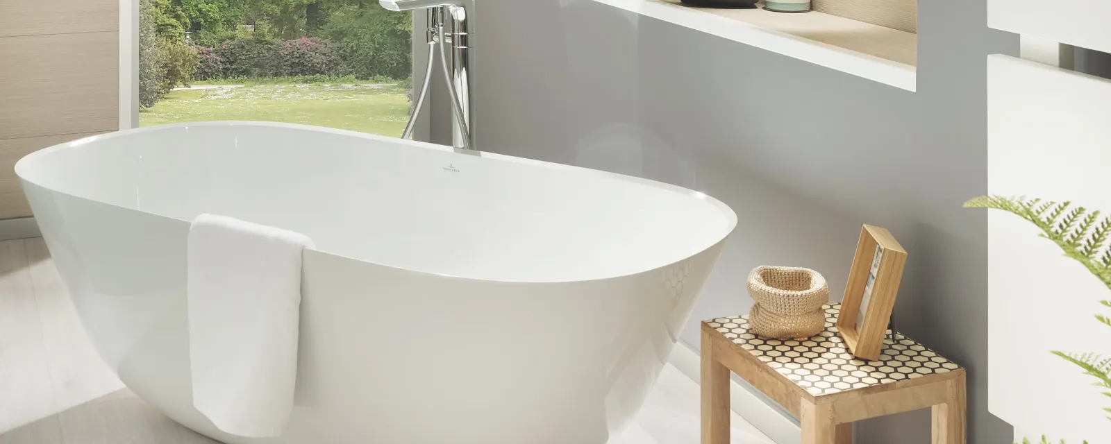 Theano Badewanne in einem modernen Badezimmer Modernes Badezimmer mit einer weißen, freistehenden Badewanne, einem darüber hängenden Handtuch, einem Holzhocker mit Toilettenartikeln und einem Spiegel. Ein eingebautes Regal mit Pflanzen und ein großes Fenster mit Blick ins Grüne ist im Hintergrund zu sehen.