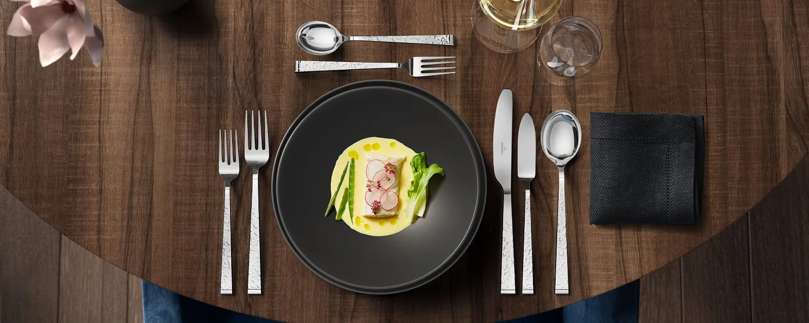 Une table élégamment dressée avec une entrée sur une table ronde sombre, accompagnée d’un verre de vin blanc et d’un ensemble de couverts de table Blacksmith.