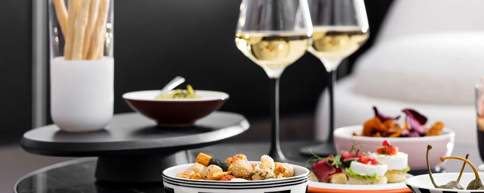 Une table noire avec des coupelles La petite Boule remplies d’amuse-bouches et de verres à vin blanc Manufacture.