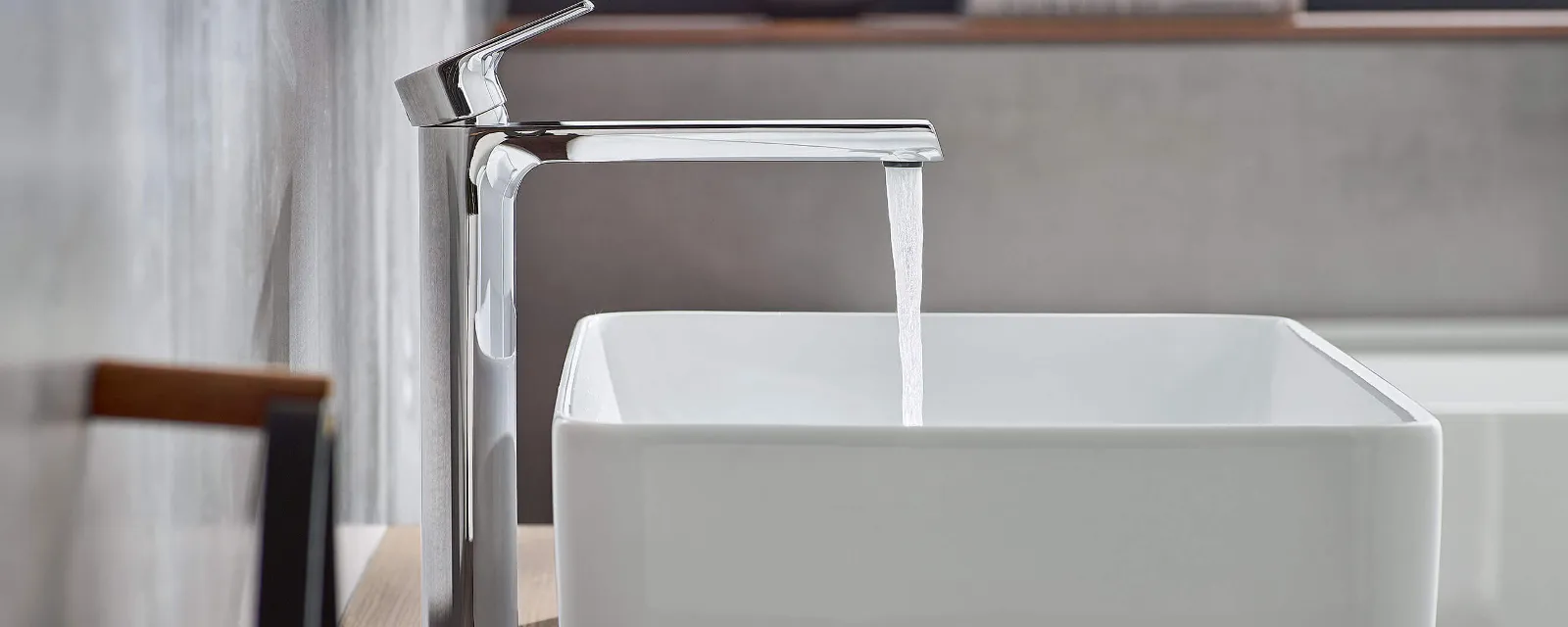 Liberty washbasin tap with a Collaro washbasin A modern chrome tap from which water is flowing into a rectangular white washbasin on a wooden shelf in a minimalist bathroom with a window in the background.