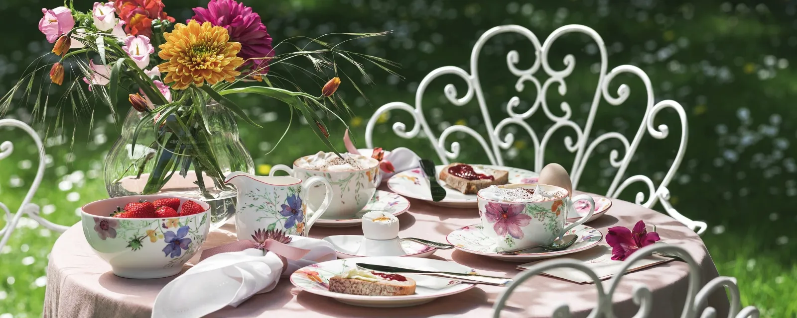 A Mariefleur garden table set from Villeroy & Boch consists of porcelain with a floral pattern, a vase full of colorful flowers, and plates decorated with desserts and strawberries. Ornate white chairs surround the table against a backdrop of lush green grass.