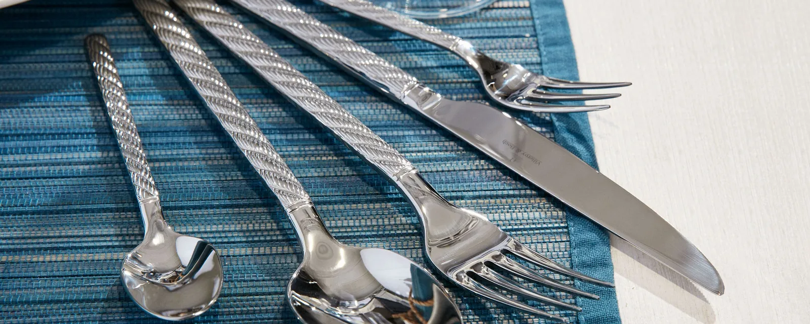 Un ensemble de couverts de table Montauk argentés disposés sur un set de table tissé bleu à côté d’une assiette blanche et d’un verre.