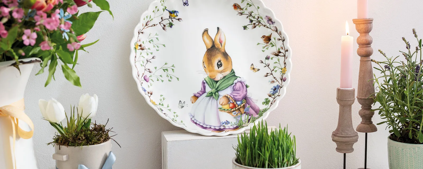 The Spring Fantasy decorative plate by Villeroy & Boch with a bunny motif is displayed on a shelf, surrounded by flowers, candles, and a wooden egg.