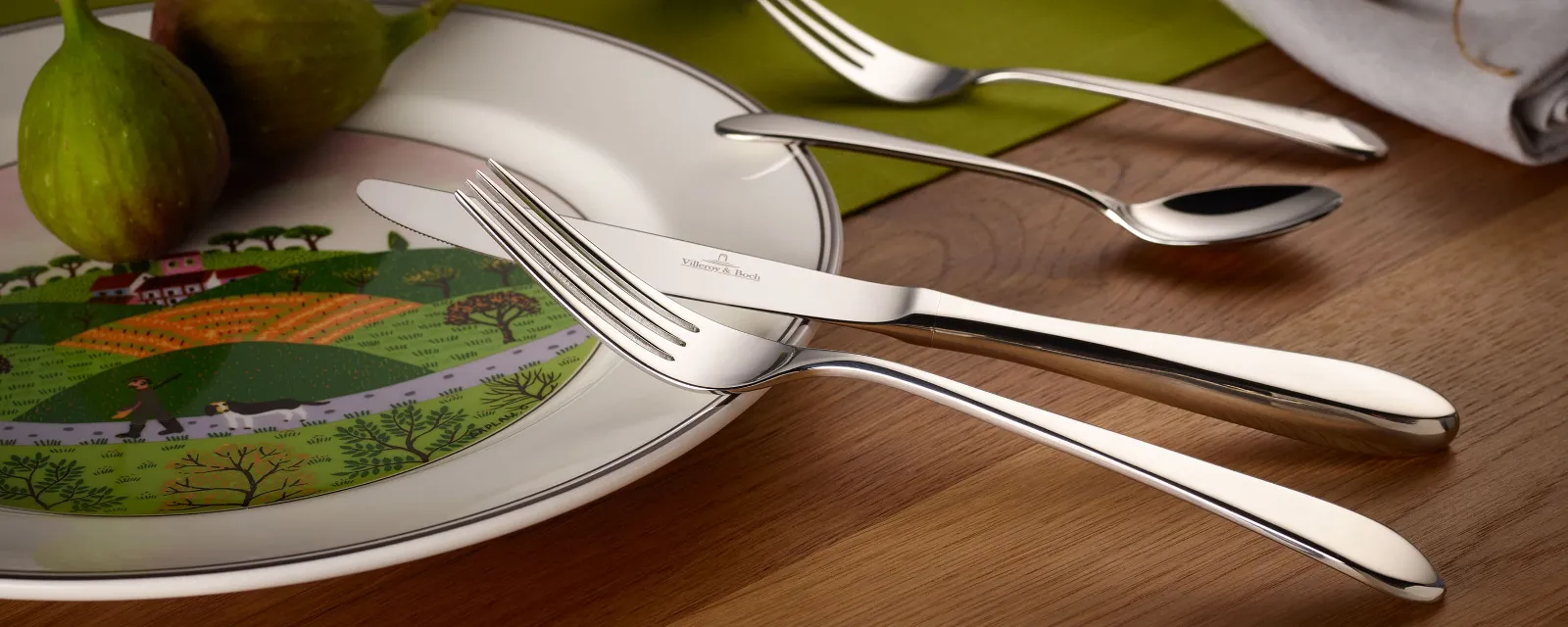 A white plate with a landscape motif, two figs, and a Sereno flatware set are arranged on a wooden table. 