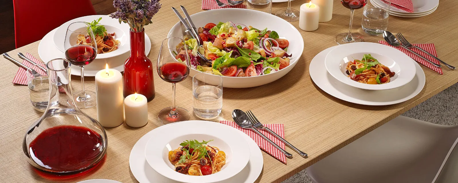 A dining table with For me plates full of salad, pasta, glasses of red wine, candles, and a carafe of red wine.