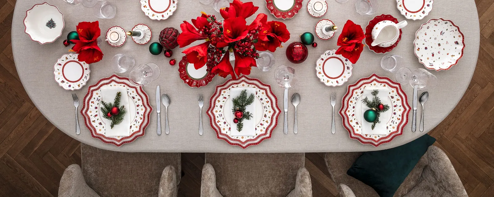 A festively decorated dining table for eight people with Villeroy & Boch Toy’s Delight red and white plates, arranged cutlery, red flowers, Christmas decorations and a grey tablecloth in a cosy dining room with grey chairs.