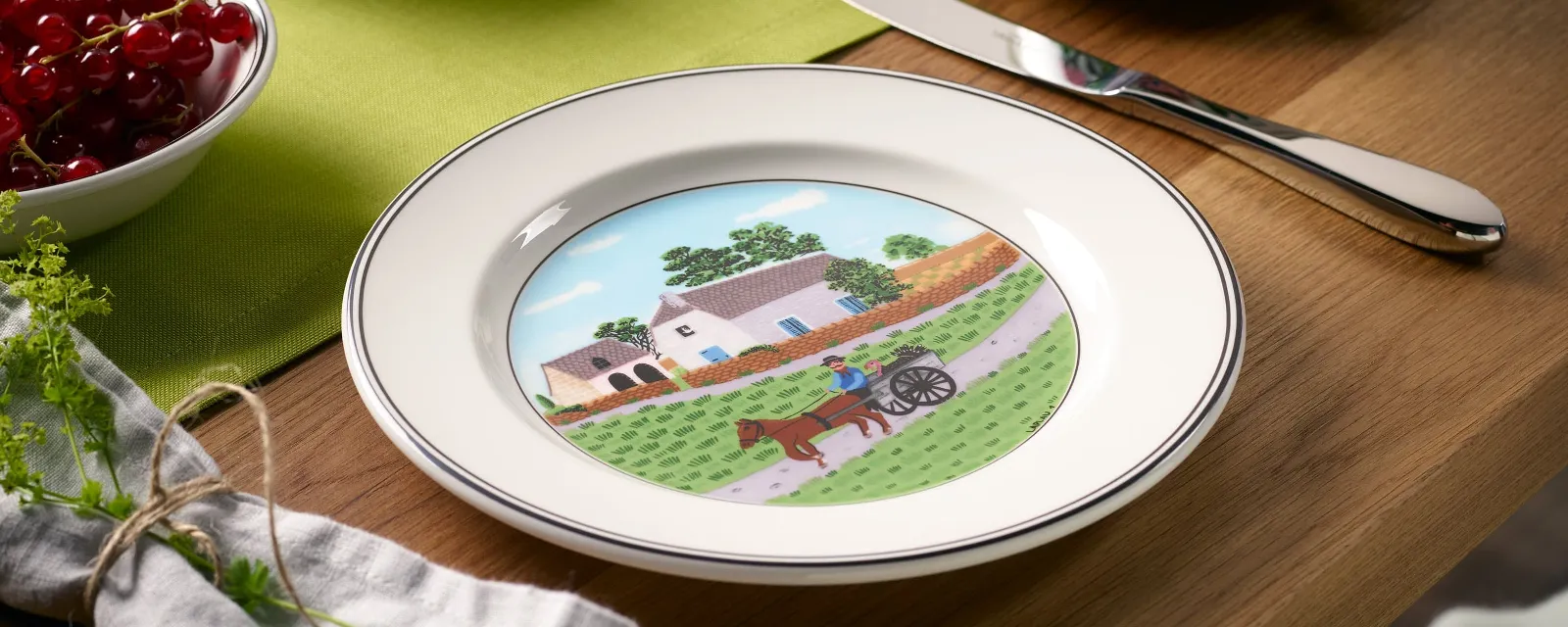 On the table is a decorative plate from Villeroy & Boch Design Naif, which depicts a picturesque landscape with a house, trees, and a horse-drawn carriage. The accompanying items include a knife, a cup, a bowl of red berries, and a green cloth.