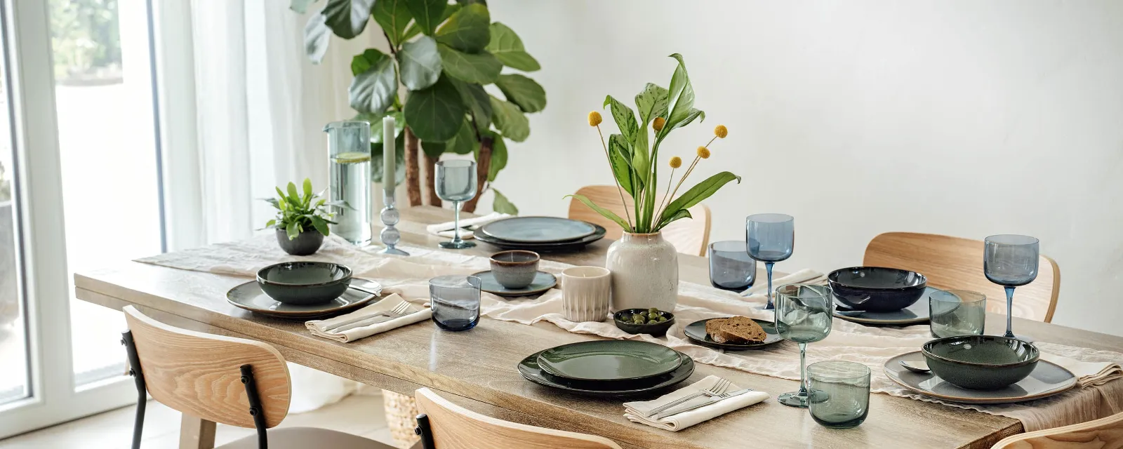 Le set de table couvert avec différents éléments de la collection Lave de Villeroy & Boch Le set de table pour la salle à manger, avec différents éléments de la collection Lave de Villeroy & Boch en vert et gris, accentués par des verres et des serviettes bleus. Un vase avec des fleurs jaunes et une petite plante en pot assurent la décoration, tandis qu'une grande plante est placée près de la fenêtre.
