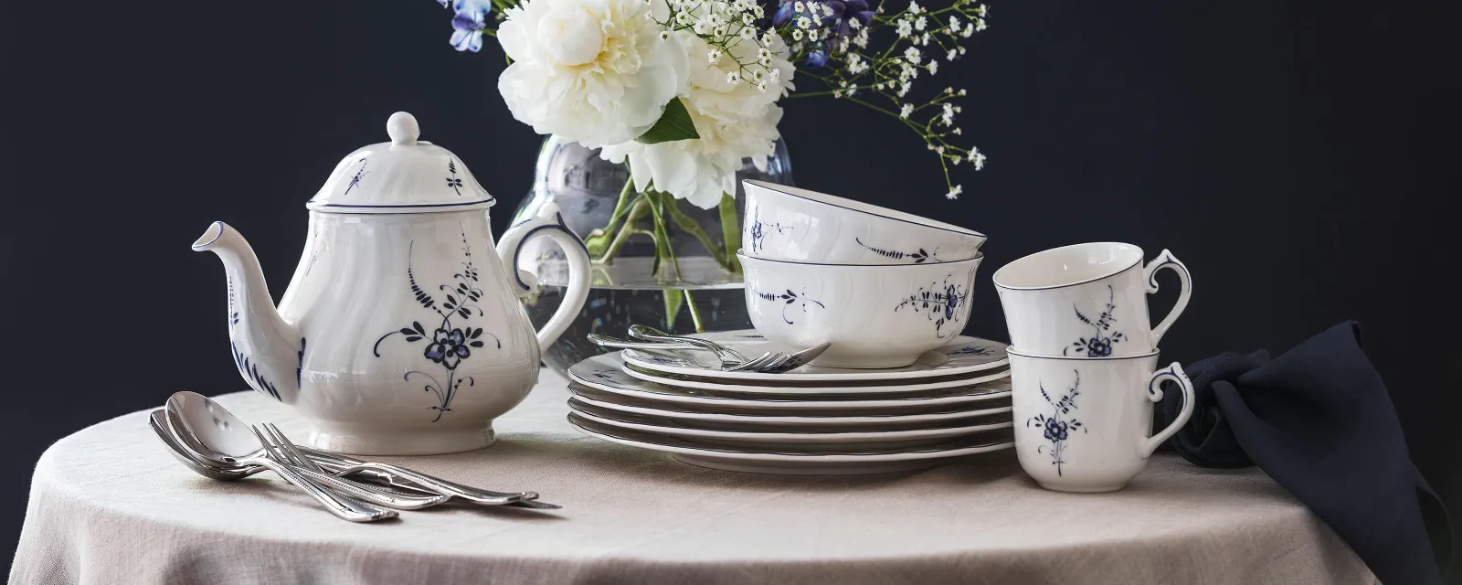 Un service en porcelaine Vieux Luxembourg avec un motif floral bleu et blanc, composé d’une théière, de tasses et d’assiettes sur une table ronde, avec en arrière-plan un vase avec des fleurs blanches et violettes.