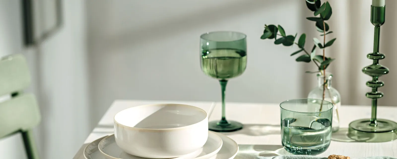 A dining table setting with white Crafted plates, green glasses from like Glass and bowls, along with green napkins with gold holders and green leaf decoration.