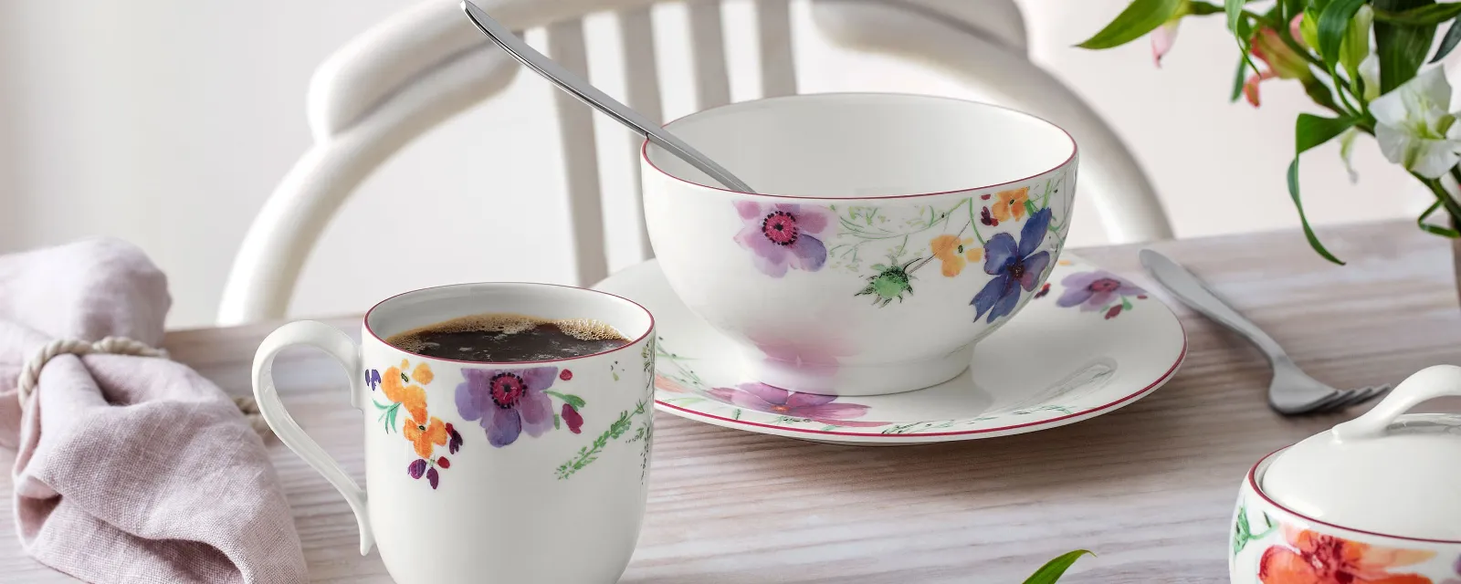 The Mariefleur collection by Villeroy & Boch includes a mug, a bowl, and a saucer with a floral pattern on a light wooden table, accompanied by a napkin, a spoon, and a small floral arrangement.