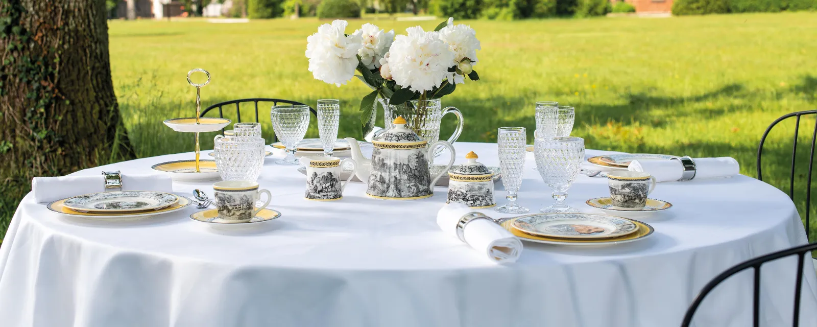 Un coin repas élégant en plein air avec une table dressée de façon raffinée Audun et des verres Boston sur une nappe blanche devant un château historique par une journée ensoleillée.