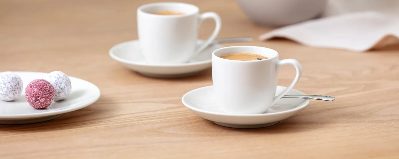 Deux tasses ForMe blanches de Villeroy & Boch sur des soucoupes avec cuillères sur une table en bois. Deux tasses ForMe blanches de Villeroy & Boch sur des soucoupes avec cuillères sur une table en bois. En arrière-plan, une assiette de truffes multicolores et un vase de fleurs roses et blanches.