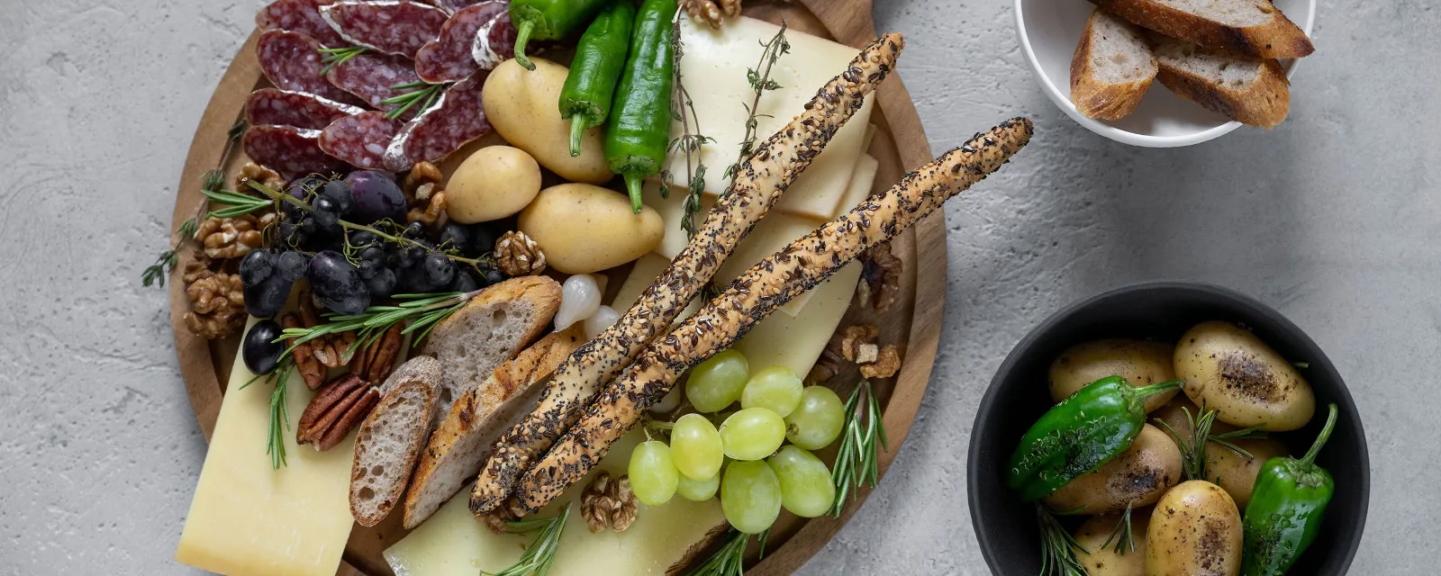 Cheese, salami, grapes, nuts, potatoes, peppers and grissini are elegantly arranged on a Villeroy & Boch Artesano wooden board. Next to this is a bowl of sliced bread and another bowl of potatoes and peppers. Cheese, salami, grapes, nuts, potatoes, peppers and grissini are elegantly arranged. 