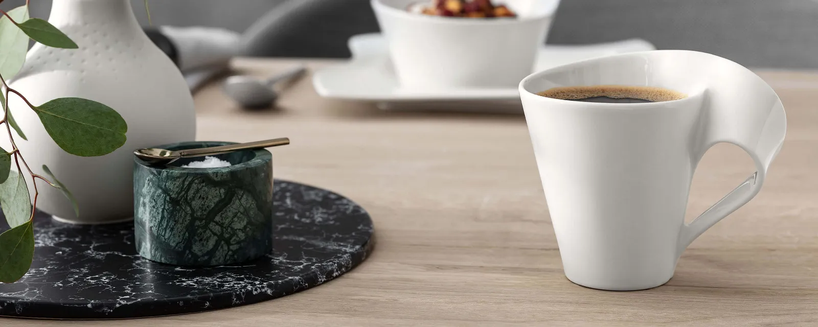 Un mug NewWave avec un vase de fleurs Une tasse à café blanche moderne avec un design d’anse unique, placée à côté d’un petit récipient et d’un vase avec des feuilles vertes sur une table en bois.