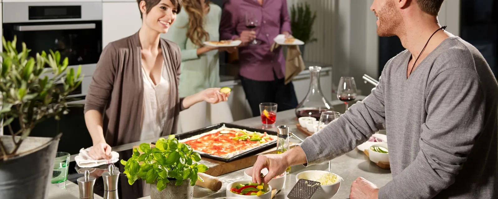 Due persone preparano pizza e cibo fresco su un'isola da cucina Kensington di Villeroy & Boch. Sullo sfondo, altre due persone stanno bevendo e chiacchierando.