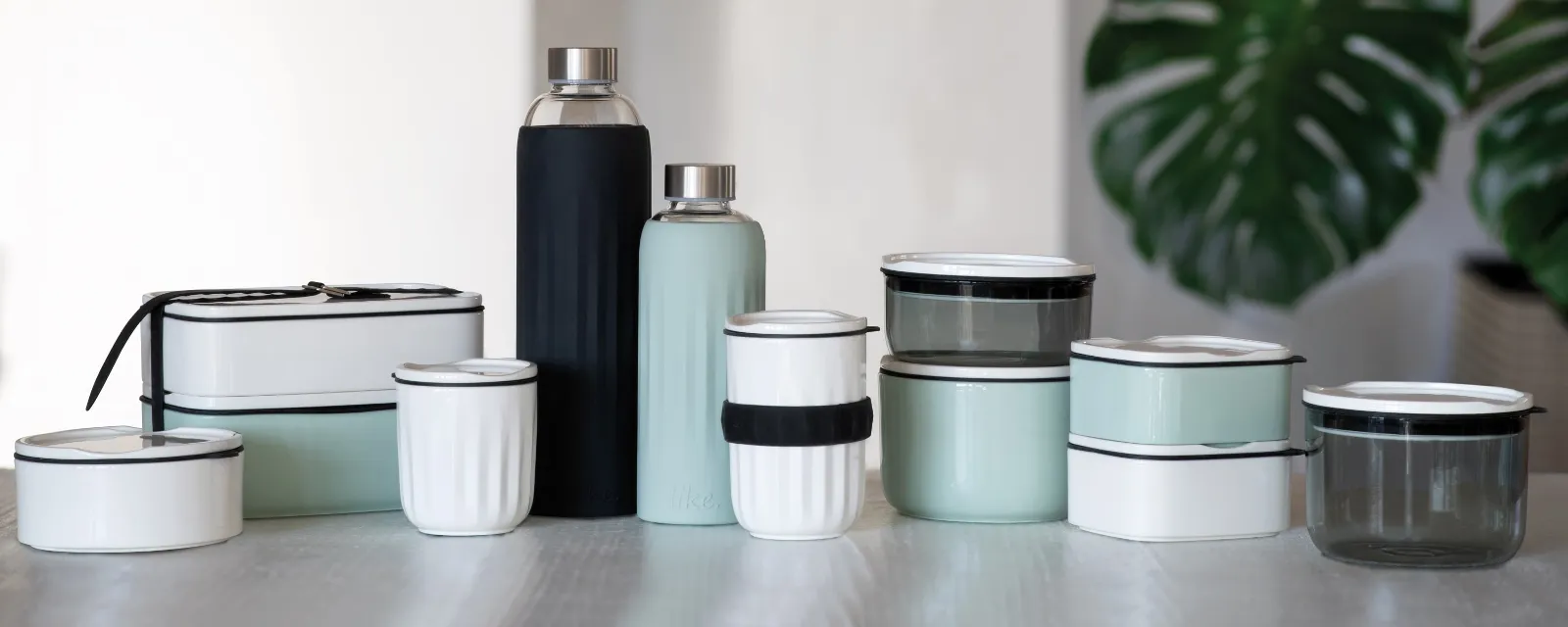 A set of reusable containers and bottles “To go and To stay” from Villeroy & Boch in different sizes and colors, arranged on a table with a potted plant in the background.