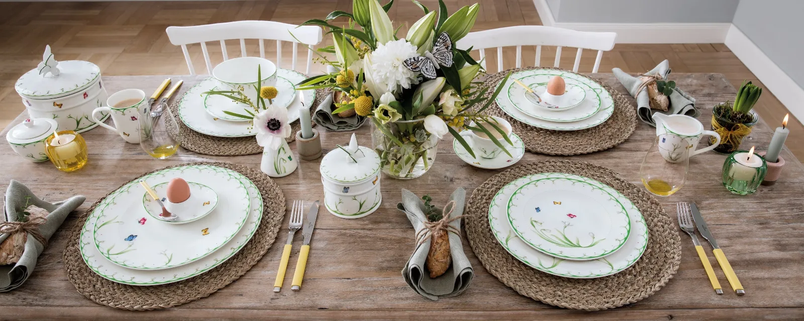 Een tafelservies "Colourful Spring" voor vier personen van Villeroy & Boch bestaande uit porselein met een bloemmotief, bestek met gele handgrepen, geweven placemats en een bloemstuk. Elk couvert bevat een ei in een decoratief kopje.