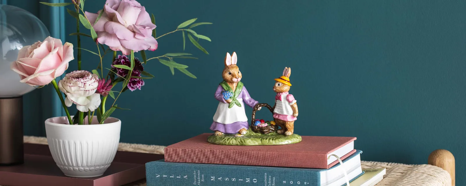 Two decorative bunny figurines from the Bunny Tales collection by Villeroy & Boch on stacked books on a wicker bench, next to a vase with ranunculus and roses.