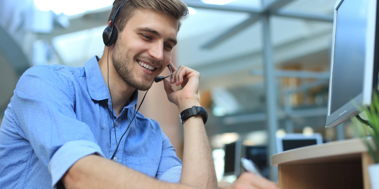 Lächelnder Mann mit Headset, der in einer Büroumgebung an einem Computer arbeitet.