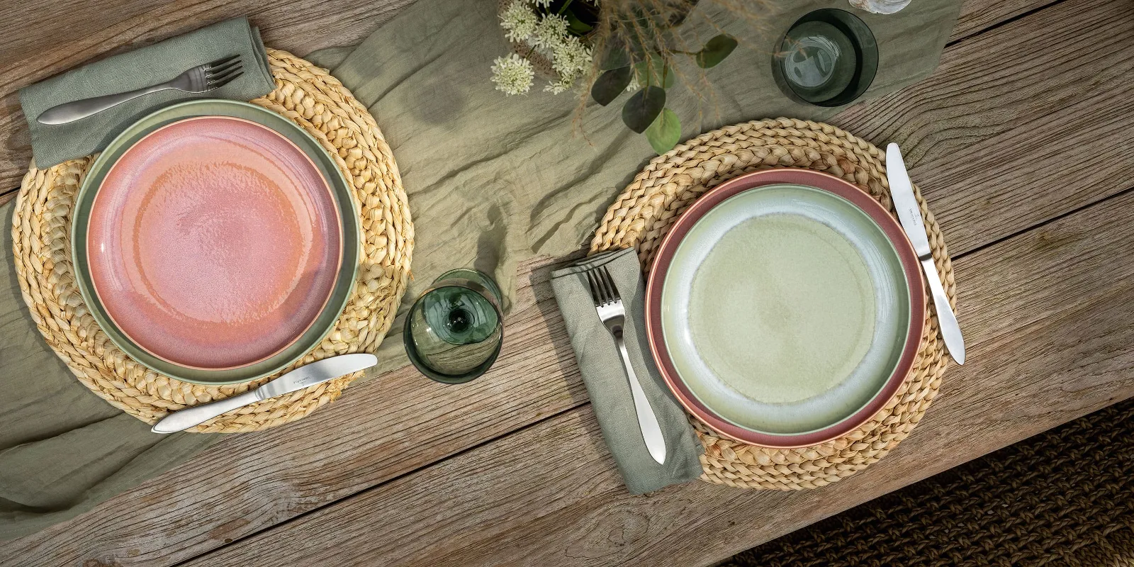 Two Perlemor place settings on a wooden table with round woven placemats, flatware, a candle, flowers, and green plants.