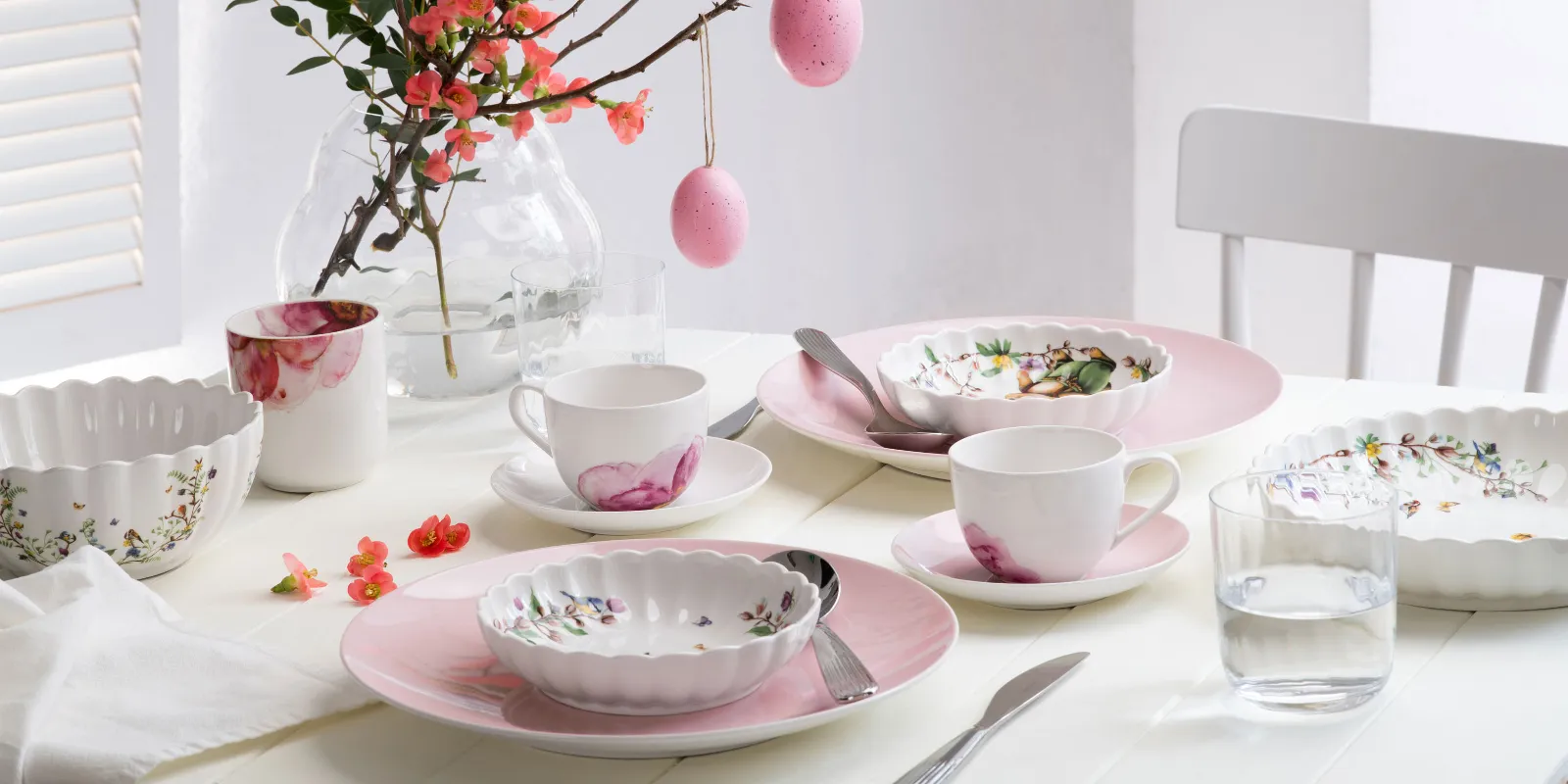 Een heldere, lenteachtige eettafel gedekt met pastelkleurig servies uit de Rose Garden en Spring Fantasy collecties, versierd met bloemmotieven en paaseieren hangend aan een tak als middelpunt.