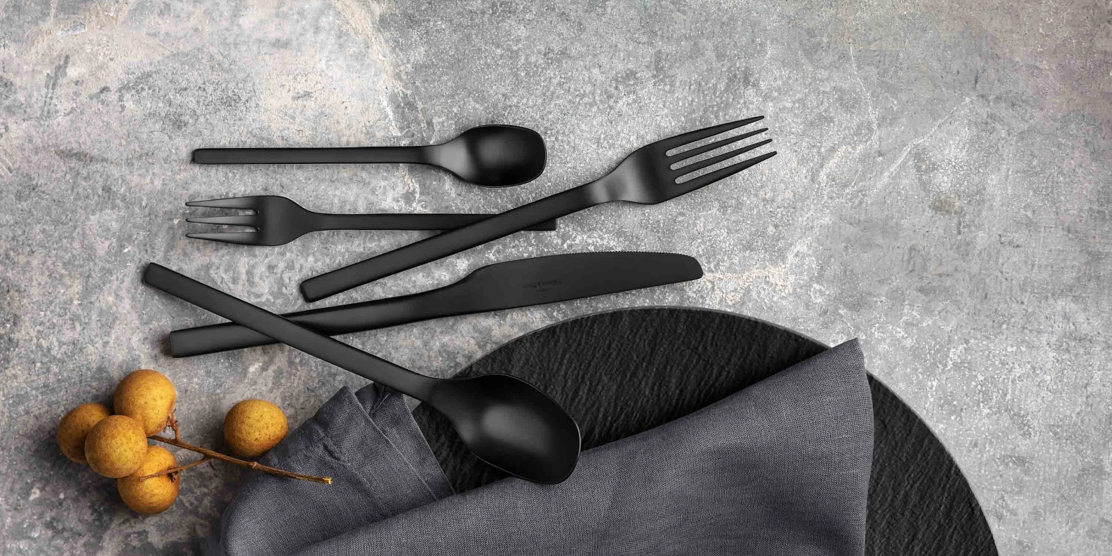 Black Manufacture flatware set with fabric napkin and decorative berries on a textured gray surface.