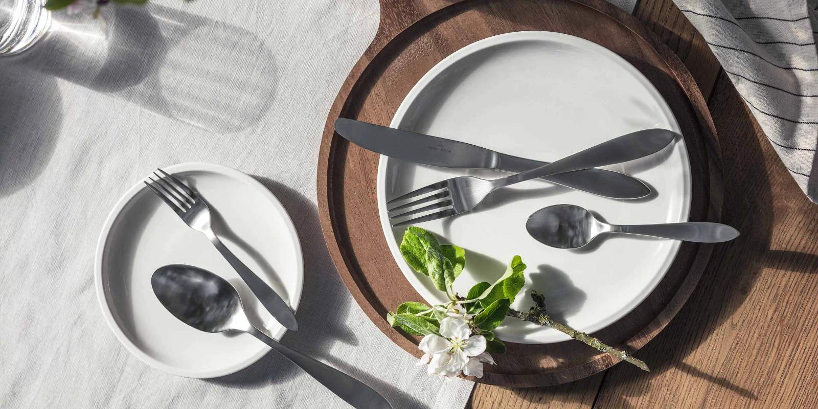 Elegante tafeldecoratie met eenvoudige witte borden en Arthur Brushed roestvrijstalen bestek, geaccentueerd door natuurlijk licht en een takje bloemen.