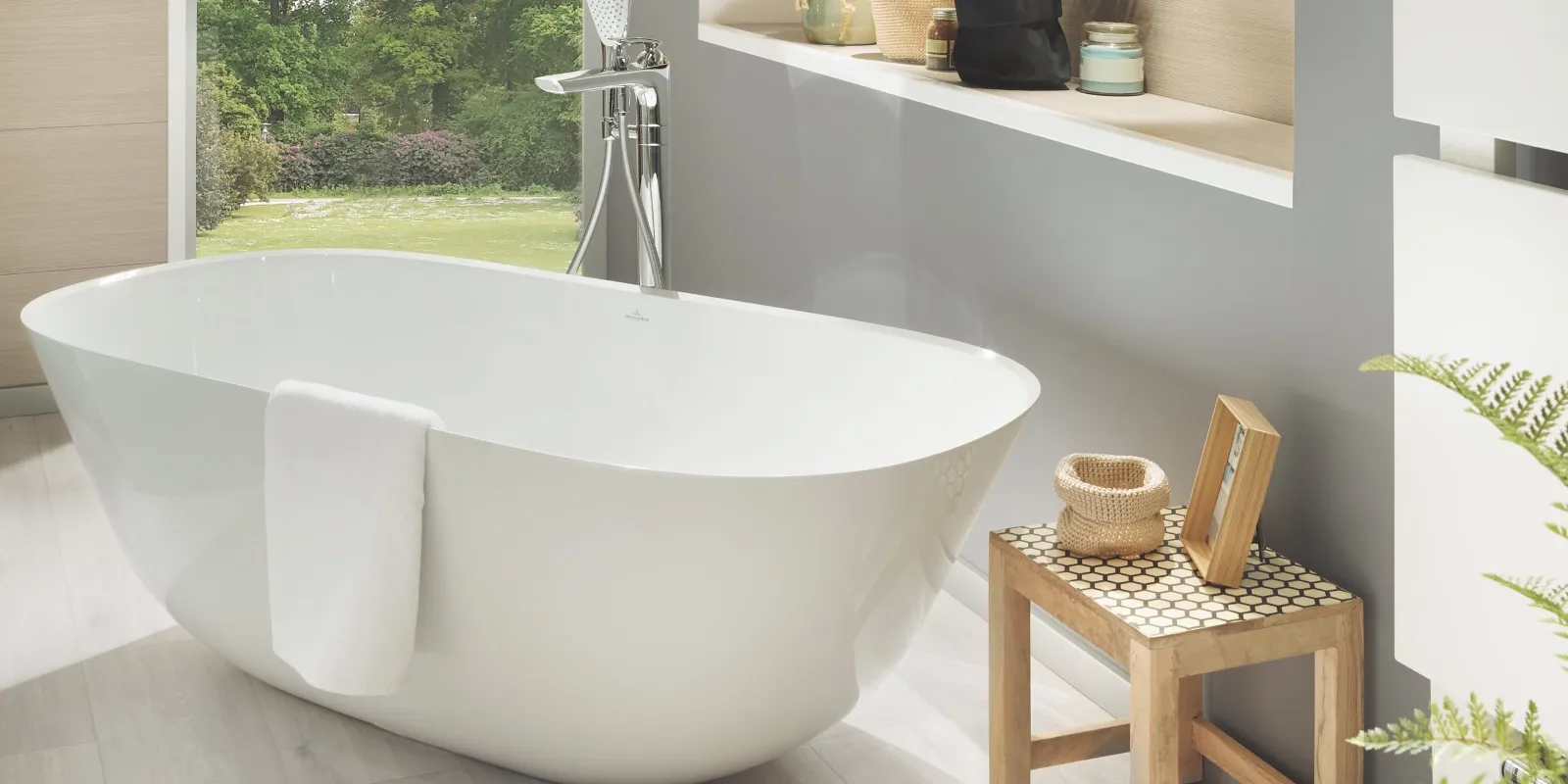 Bagno moderno con vasca bianca free-standing, asciugamano appeso, sgabello in legno con articoli da bagno e specchio. Sullo sfondo si intravedono uno scaffale a muro con piante e un'ampia finestra con vista sul verde.