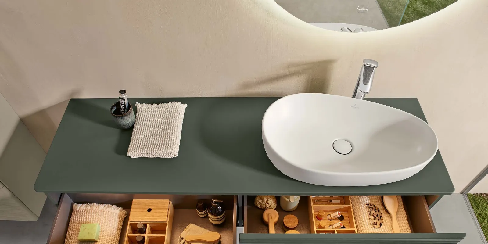 Une console d’un vert naturel dans la salle de bains avec deux tiroirs ouverts et remplis, surmontée d’une vasque blanche Antao de Villeroy & Boch.