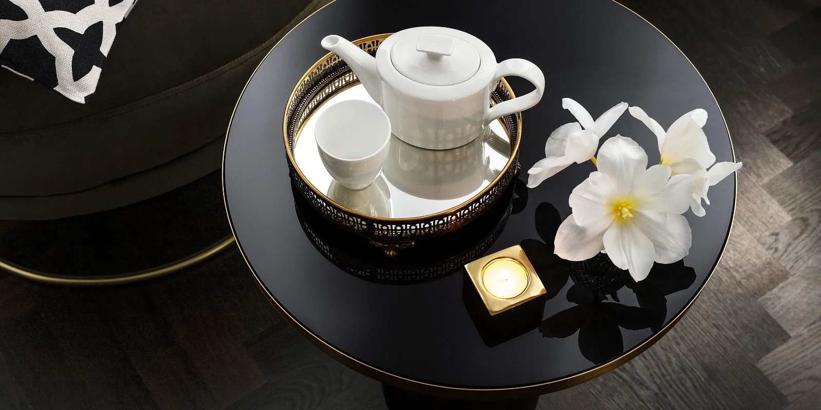 A white MetroChic teapot with a matching cup, two white flowers and a lit candle on a round black table with a patterned cushion in the background.