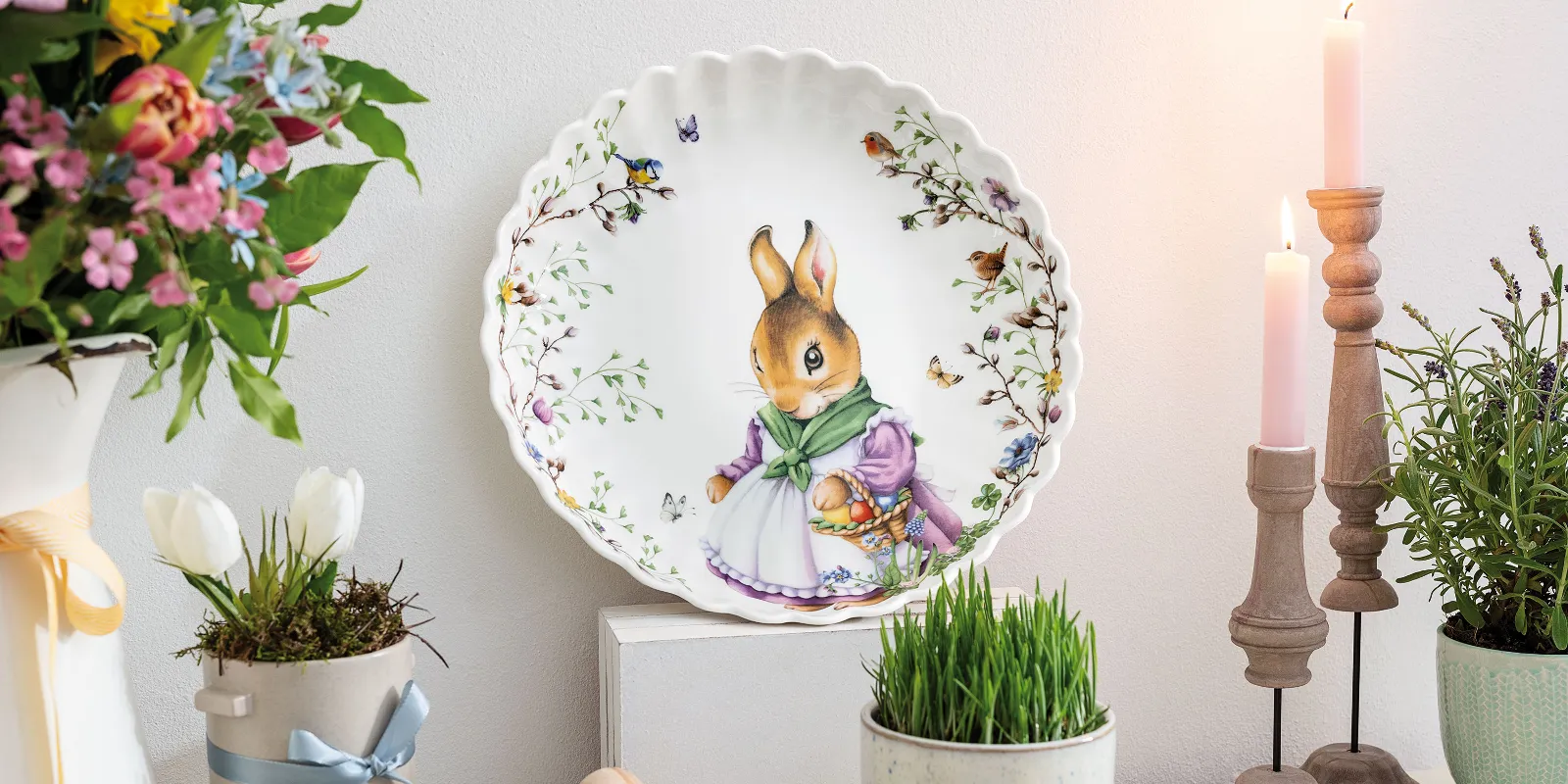 The Spring Fantasy decorative plate by Villeroy & Boch with a bunny motif is displayed on a shelf, surrounded by flowers, candles, and a wooden egg.