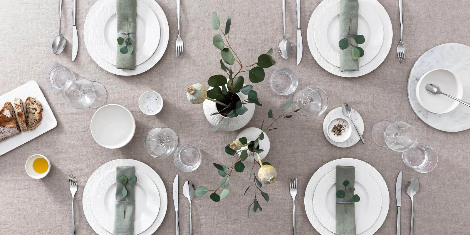 Modern dining table for six people with white Manufacture plates, cutlery and green floral decoration, seen from above.