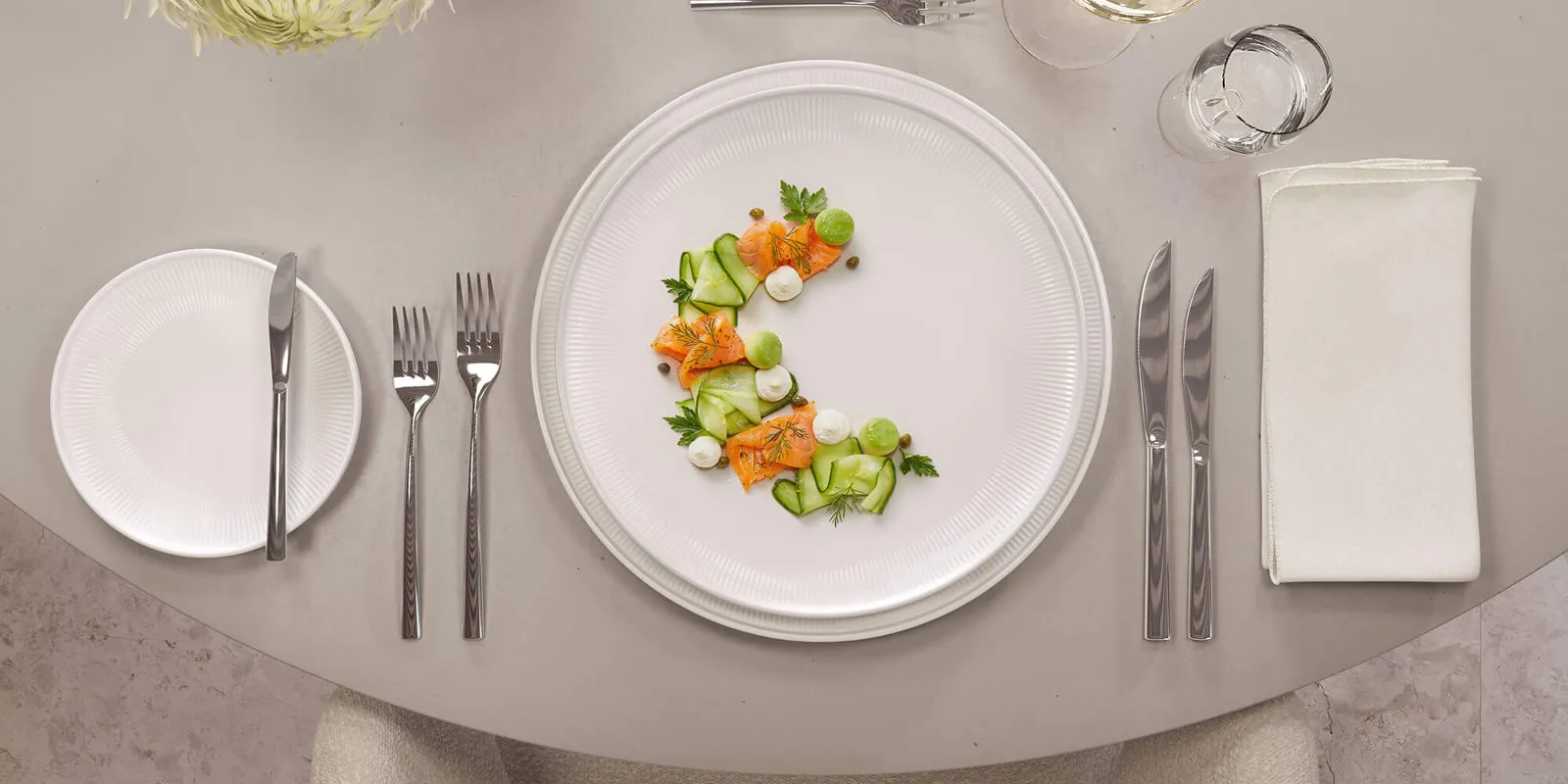 Elegant dining table with salmon and cucumber salad on Afina plates, accompanied by wine in La Divina glasses and Piemont flatware.