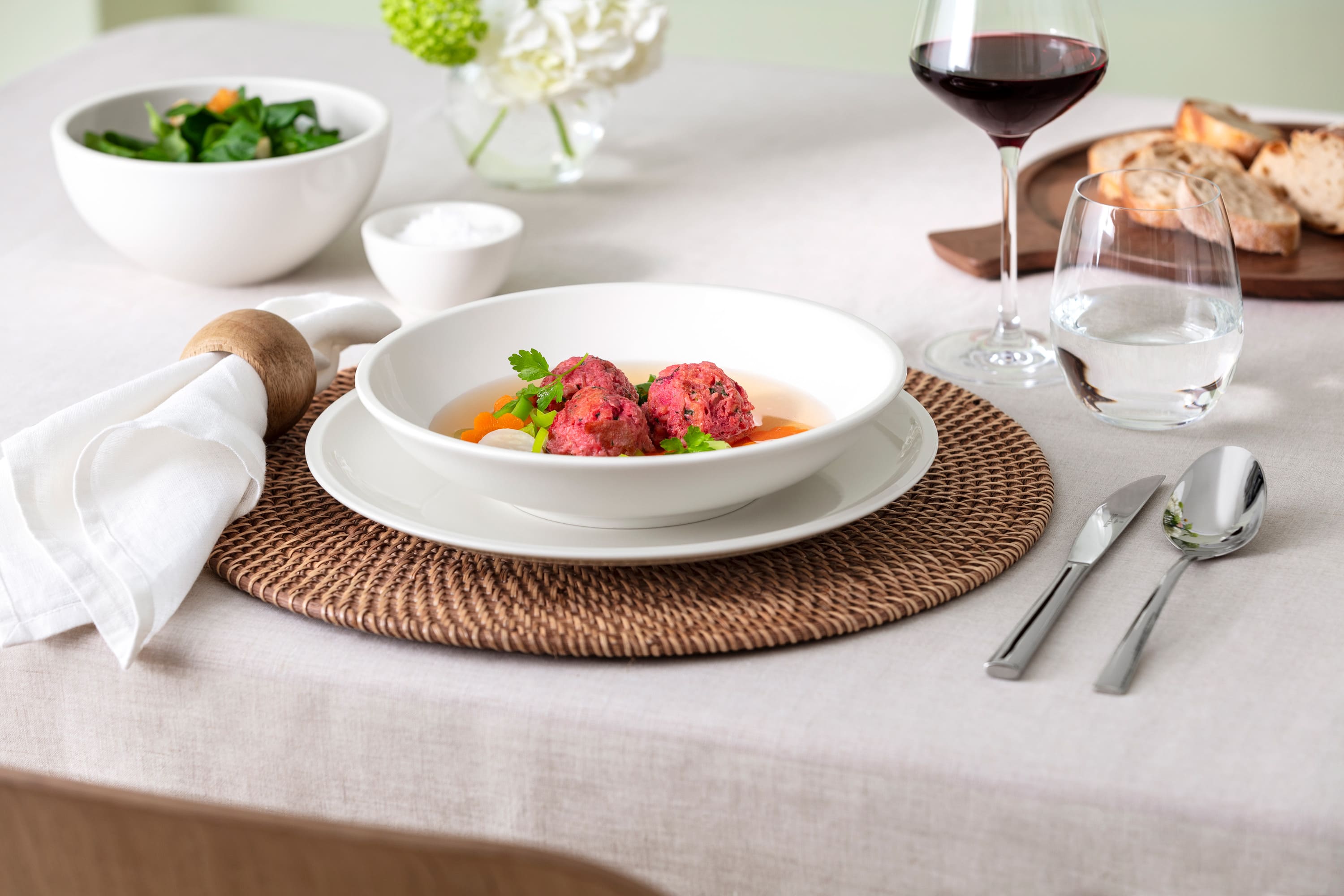 A set table with an Artesano plate featuring beetroot balls, carrots, and some greens. A glass of red wine, water, a salad bowl, bread, and cutlery are arranged on a woven placemat and a white tablecloth.