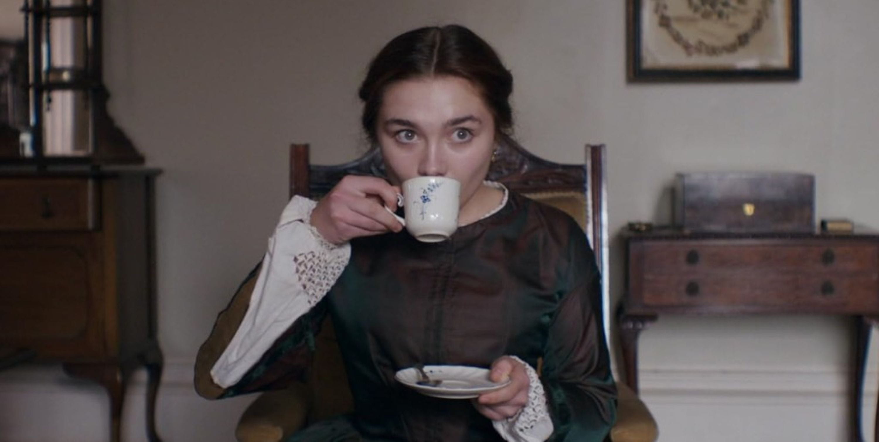 lady-mcbeth-as-seen-on A woman in a vintage green dress sips from a teacup, seated in a wooden chair. The room has a calm, historical ambiance with a framed picture and cabinet.
